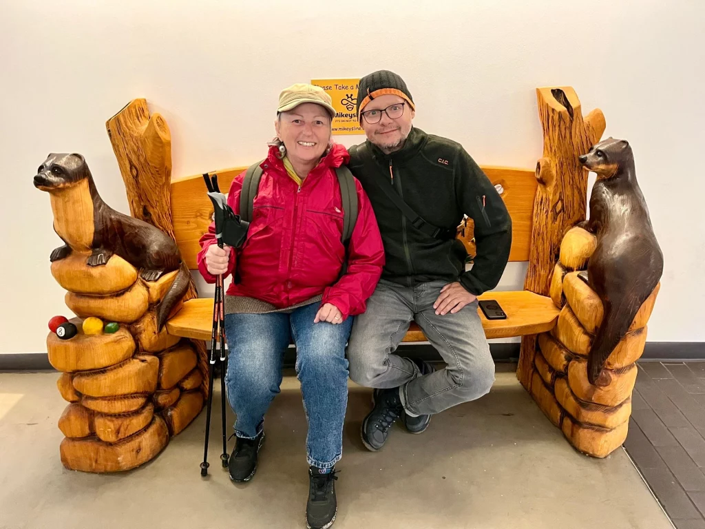 Ein Paar sitzt lächelnd auf einer rustikalen Holzbank, deren Armlehnen als detaillierte Otter-Skulpturen geschnitzt sind. Die Bank steht vor einer weißen Wand im Innenbereich.