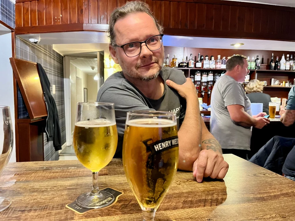 Nahaufnahme eines Mannes mit Brille an einem Holztisch in einem Pub, im Vordergrund stehen zwei gefüllte Biergläser vor einer belebten Bar-Kulisse.