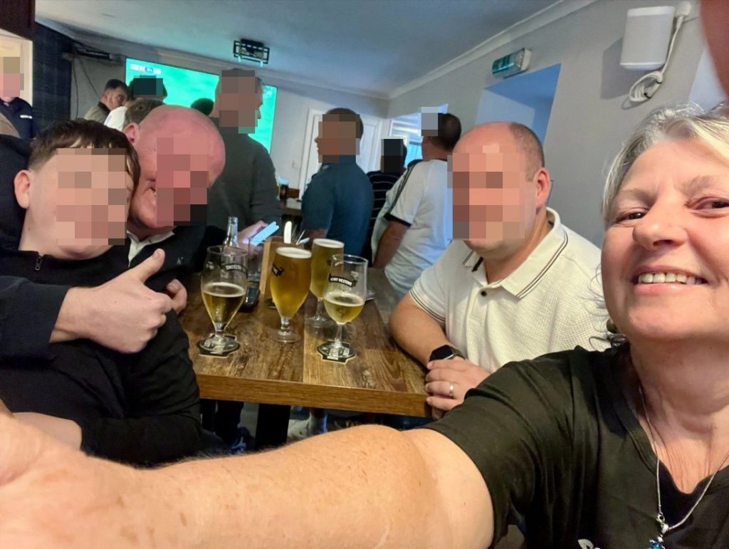 Eine Gruppe von Menschen sitzt gut gelaunt in einem Pub an einem Holztisch mit mehreren Gläsern Cidre; im Hintergrund läuft ein Fernseher.