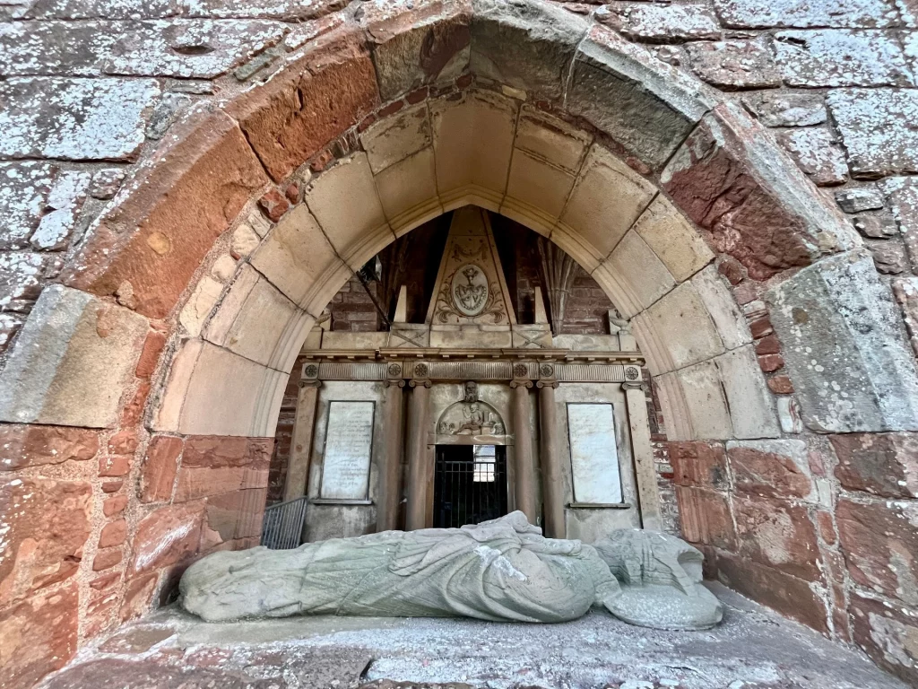 Blick durch einen Steinbogen auf ein großes Wandgrabmal mit einer liegenden, stark verwitterten Steineffigie im Vordergrund.