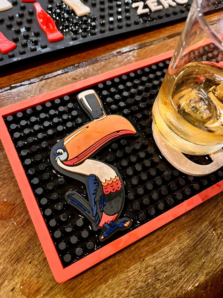 Eine rote Gummimatte auf einem Holztresen mit dem bunten Logo des Guinness-Tukans, der ein Glas Bier auf dem Schnabel balanciert. Daneben steht ein Glas mit einem goldfarbenen Getränk.