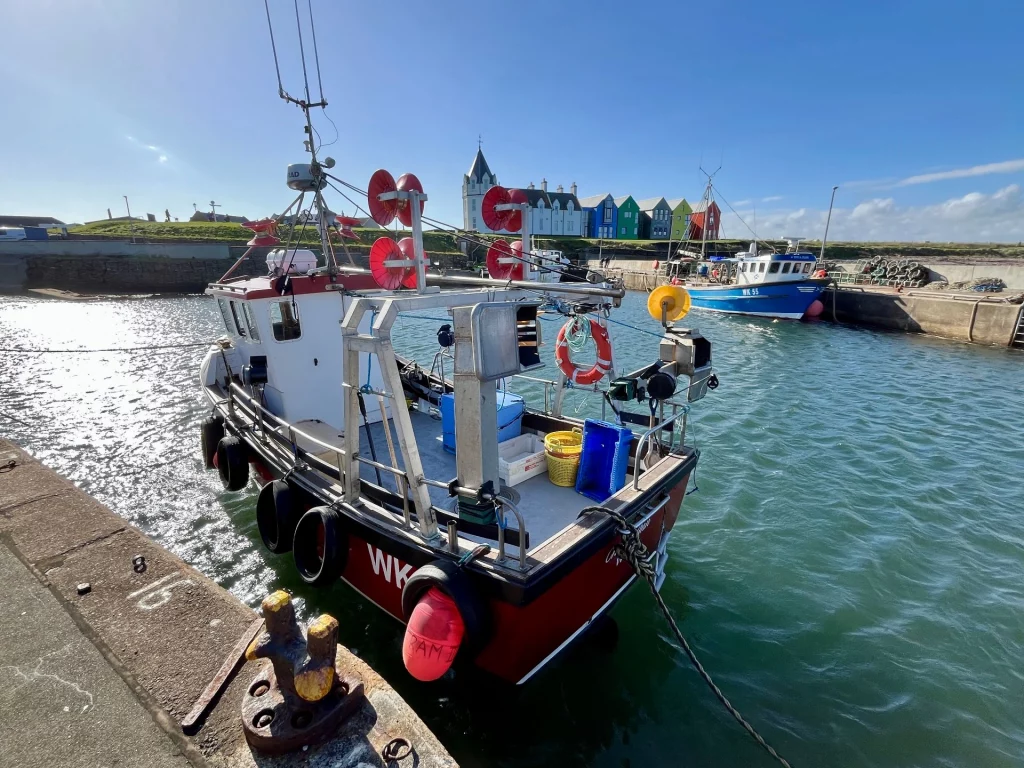 Blick vom Kai auf ein rotes Fischerboot im Vordergrund und die berühmten bunten Häuser von John o' Groats, deren idyllische Kulisse zunehmend durch die Folgen vom Klimawandel bedroht wird.