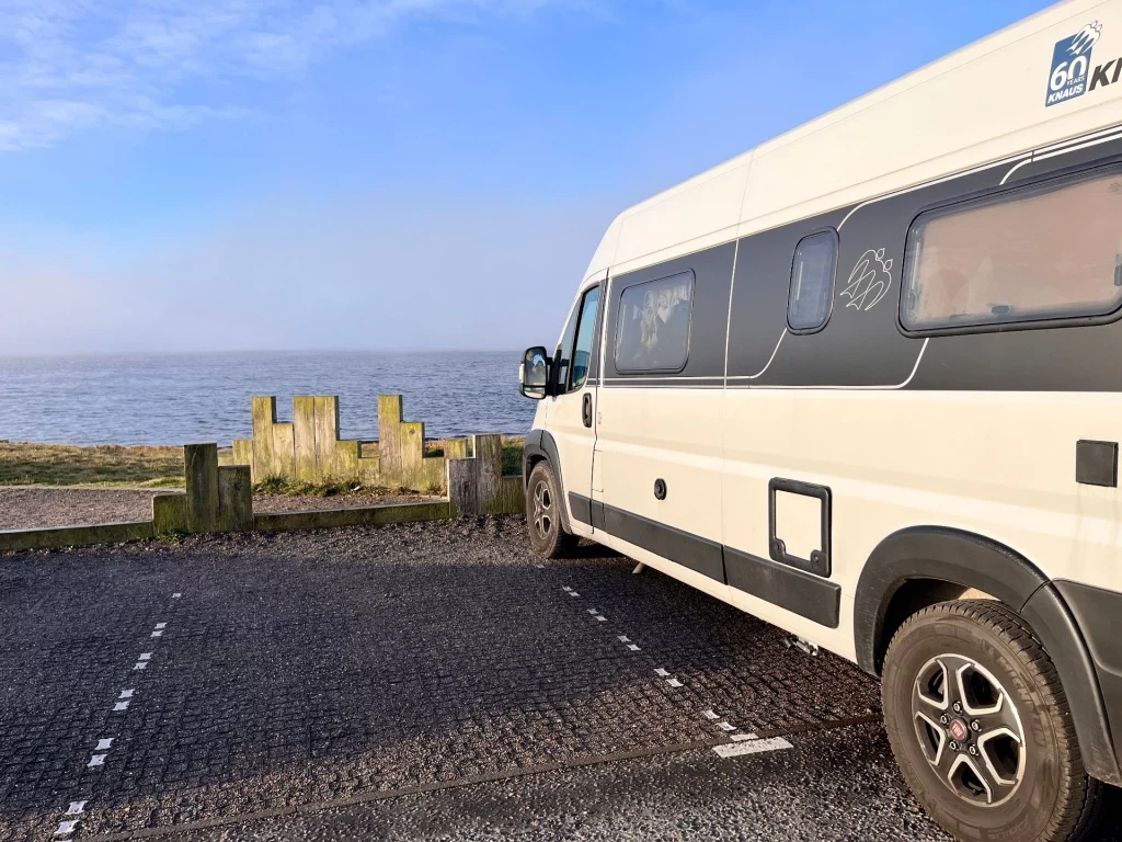 Seitenansicht eines grauen Knaus Kastenwagens, der auf einem Schotterparkplatz direkt am Meer steht; im Hintergrund ein hölzerner Wellenbrecher und ruhiges Wasser.