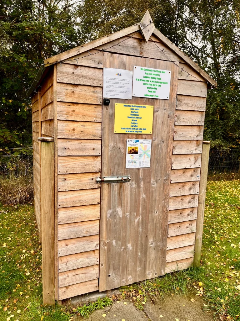 Eine kleine, wettergebräunte Holzhütte auf einer Wiese. An der Tür hängen mehrere laminierte Schilder mit Informationen zur Nutzung und Tipps für geeignete Lebensmittelspenden.