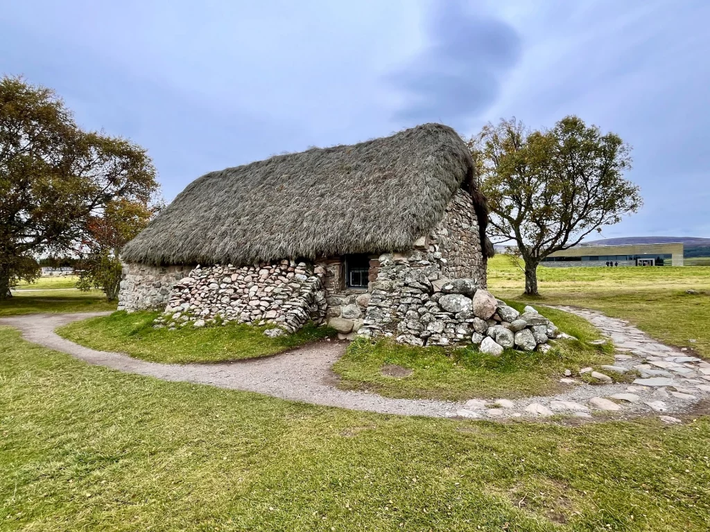 Ein kleines, historisches Steinhaus mit einem dicken, grauen Reetdach auf einer grünen Wiese. Im Hintergrund ist das moderne Besucherzentrum unter einem bewölkten Himmel zu sehen.