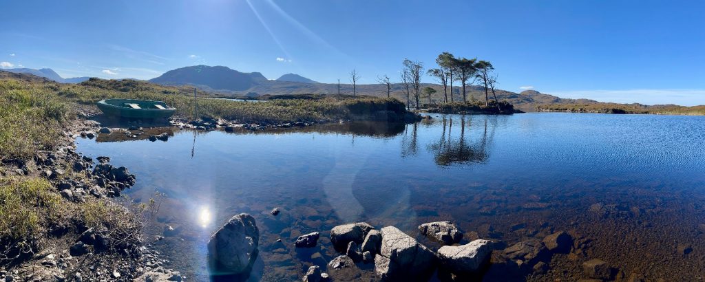 Weitwinkel-Panorama des Loch Assynt in Schottland mit einem kleinen grünen Ruderboot am felsigen Ufer und einer markanten Kieferninsel, die sich im spiegelglatten, klaren Wasser unter strahlender Sonne spiegelt.