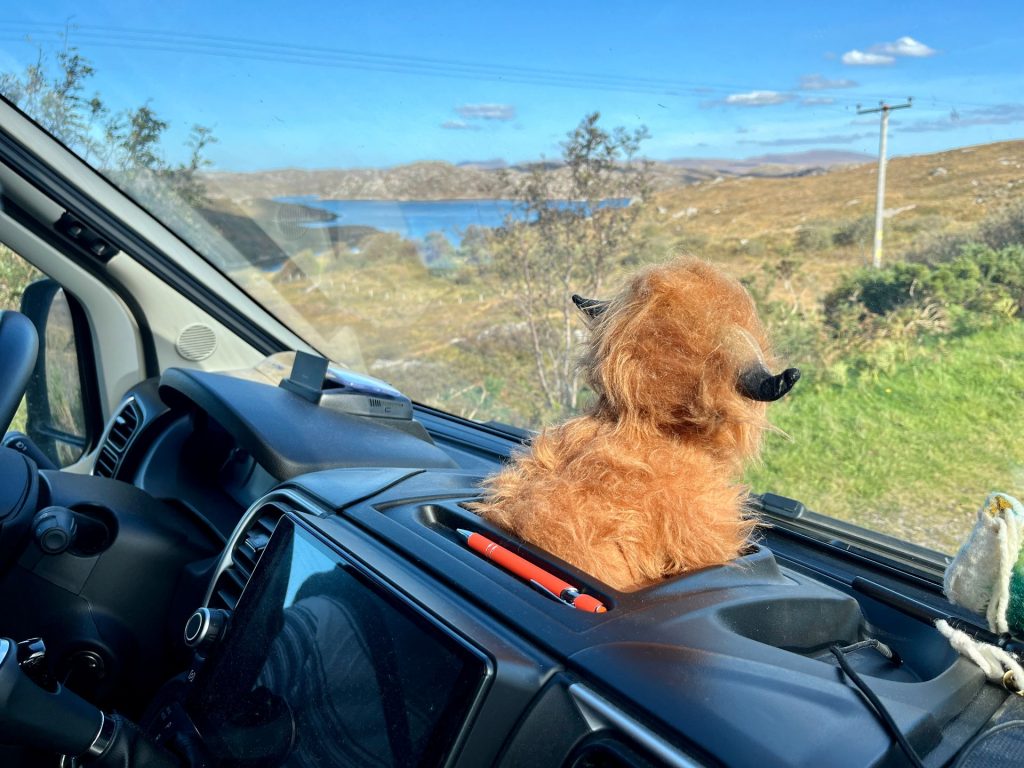 Ein kleines, plüschiges Highland-Rind namens Angus sitzt als Maskottchen auf dem Armaturenbrett des Campervans und blickt durch die Windschutzscheibe auf die vorbeiziehende Landschaft der schottischen Highlands.