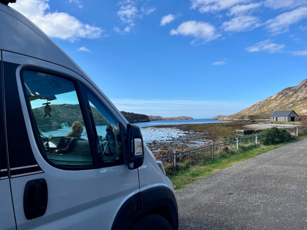 Ein silberner Knaus Campervan parkt auf einem Schotterplatz am Loch Glencoul, im Hintergrund steht das einsame „Little Cullaig Bothy“ vor einer sanften Hügelkette.