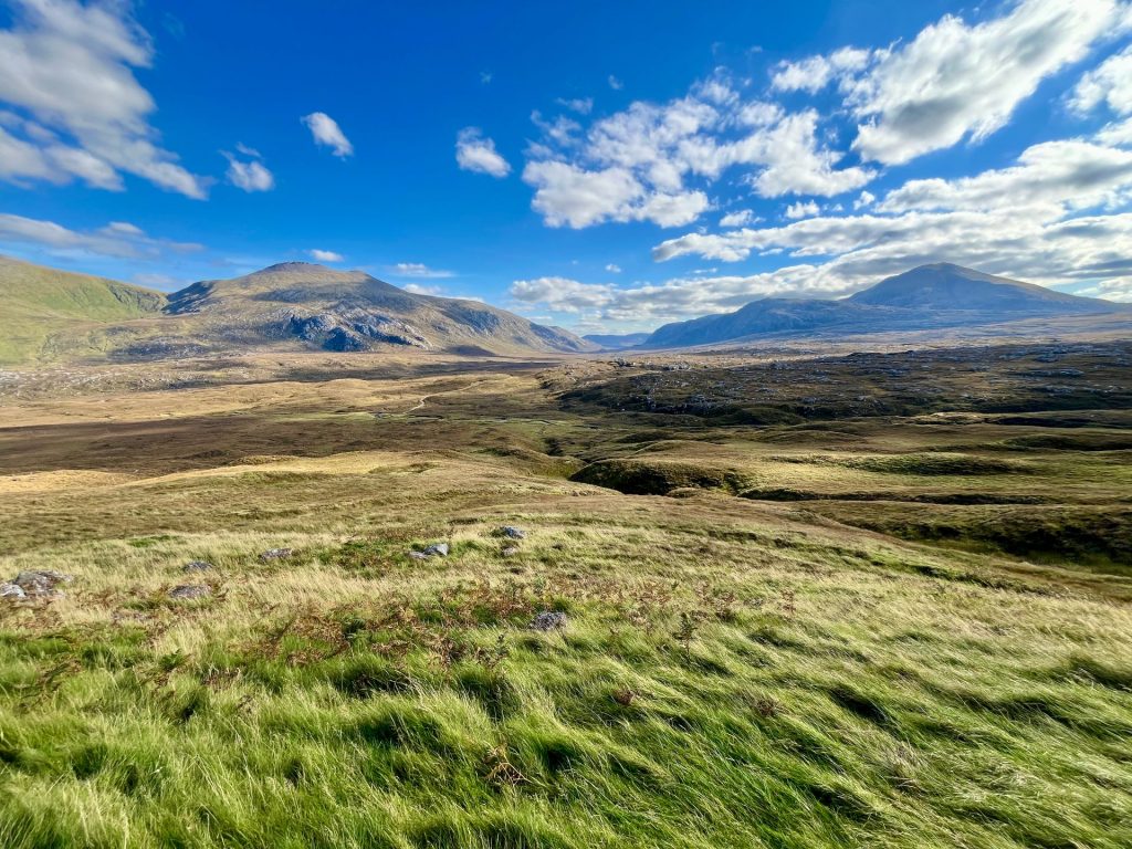 Weite, hügelige Moorlandschaft in Schottland unter einem strahlend blauen Himmel mit weißen Wolken. Im Hintergrund erheben sich sanfte, dunkle Berge, während im Vordergrund windgepeitschtes grünes Gras dominiert.