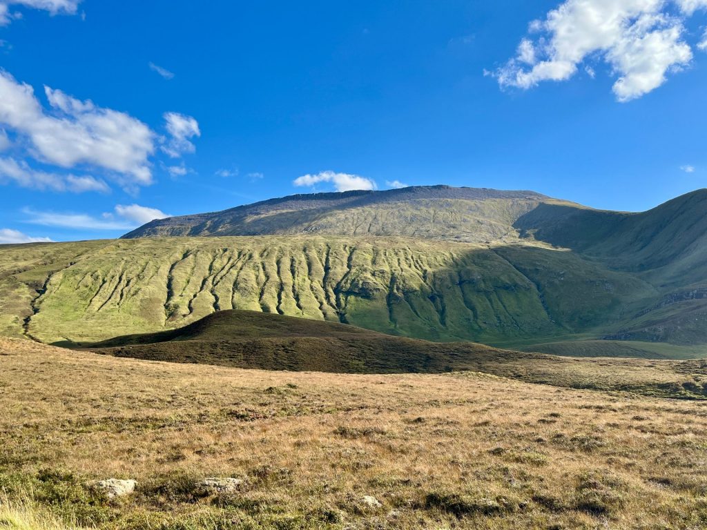 Eine markante, steile Bergflanke mit tiefen, vertikalen Furchen und sattgrünem Bewuchs. Davor erstreckt sich eine weite Ebene mit trockenem, bräunlichem Gras unter einem weiten, blauen Himmel.