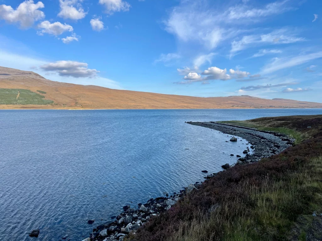 Blick entlang der felsigen Uferlinie von Loch Eriboll in Nord-Schottland mit sanften braunen Hügelketten im Hintergrund unter blauem Himmel.