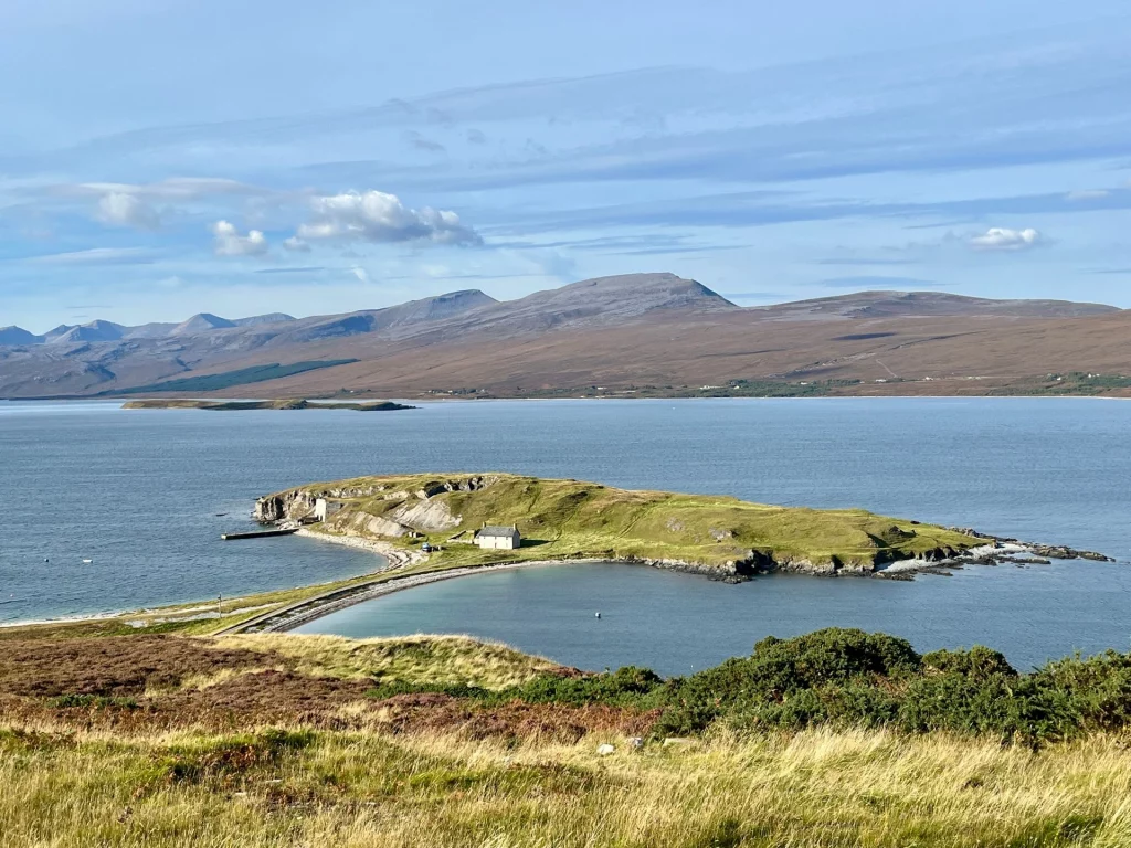 Eine weite Landschaftsaufnahme von Loch Eriboll in den schottischen Highlands unter einem leicht bewölkten, blauen Himmel. Im Mittelpunkt steht die markante Halbinsel Ard Neackie, die über einen schmalen, gebogenen Landstreifen mit dem Ufer verbunden ist. Auf der grünen Halbinsel stehen ein einsames weißes Haus und historische Kalköfen aus Stein. Im Hintergrund erstrecken sich sanfte, braun-goldene Hügelketten bis zum Horizont. Der Vordergrund zeigt eine wilde Wiese mit hohem, vom Wind gebeugtem Gras und Heidekraut. Die Atmosphäre ist friedlich, weitläufig und typisch für die nordische Küstenlandschaft.