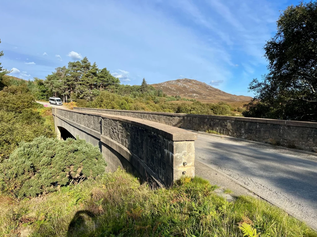 Malerische Aufnahme einer hohen Steinbogenbrücke über einem bewaldeten Tal. Im Hintergrund erheben sich herbstliche Hügel unter blauem Himmel mit Zirruswolken. Nadelbäume und buntes Laub rahmen die einsame Highland-Szenerie friedlich ein.
