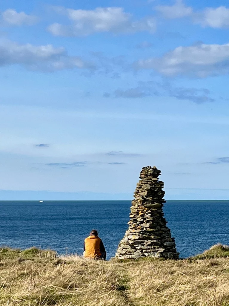 Eine Person in einer orangen Weste sitzt mit dem Rücken zum Betrachter im trockenen Gras an einer Klippe. Rechts daneben steht ein mannshoher, kunstvoll aufgetürmter stummer Wächter (Cairn) aus flachen Schieferplatten. Im Hintergrund erstreckt sich das tiefblaue Meer unter einem leicht bewölkten Himmel bis zum Horizont.