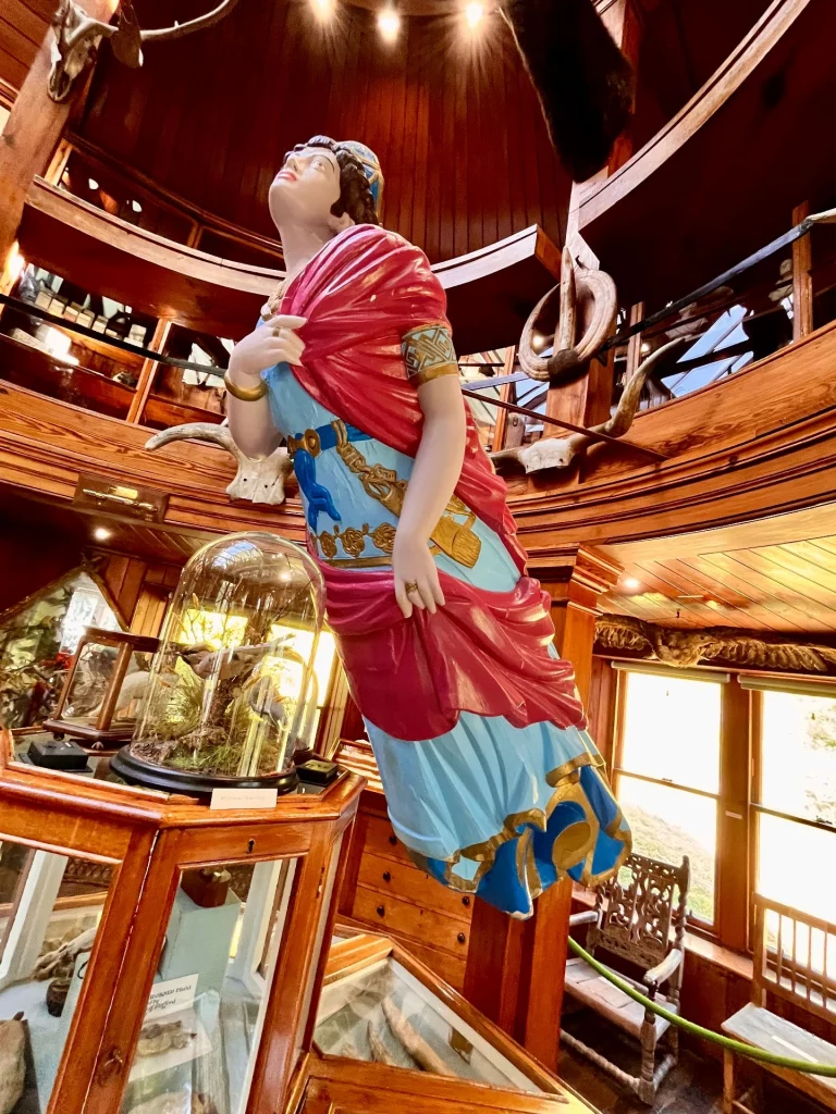 Eine farbenfroh bemalte, hölzerne Galionsfigur einer Frau in einem rot-blauen Gewand, die im prunkvollen, holzgetäfelten Innenraum des Museums ausgestellt ist.