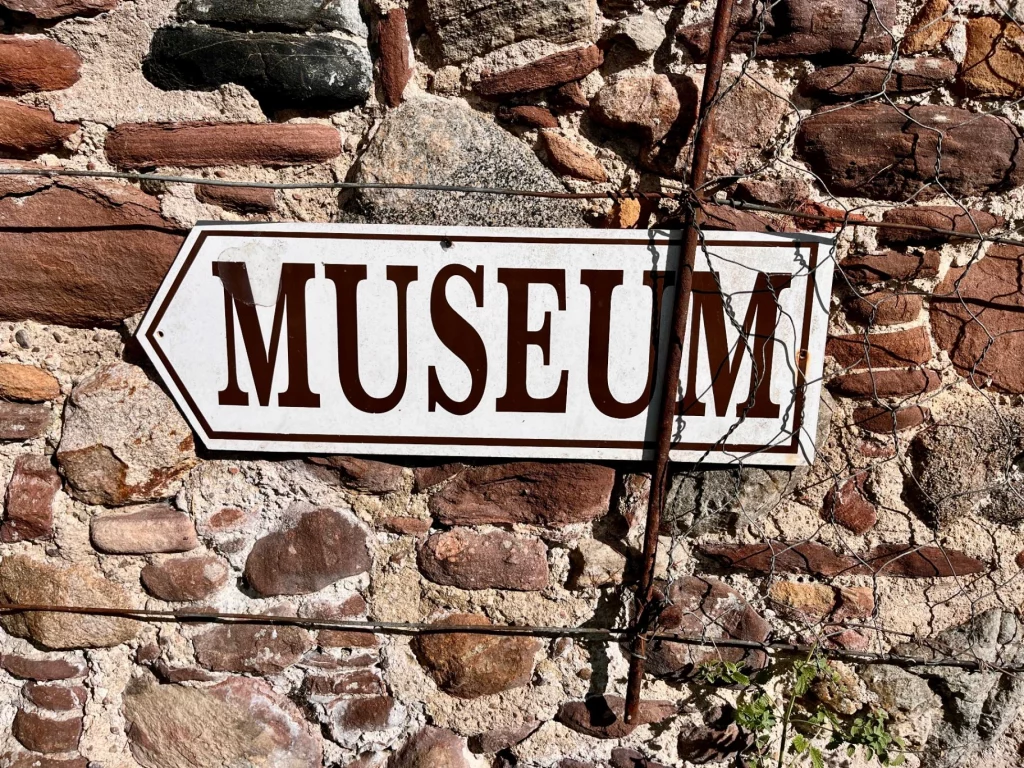 Ein weißes, pfeilförmiges Schild mit der braunen Aufschrift „MUSEUM“, das an einer rustikalen Natursteinmauer befestigt ist.