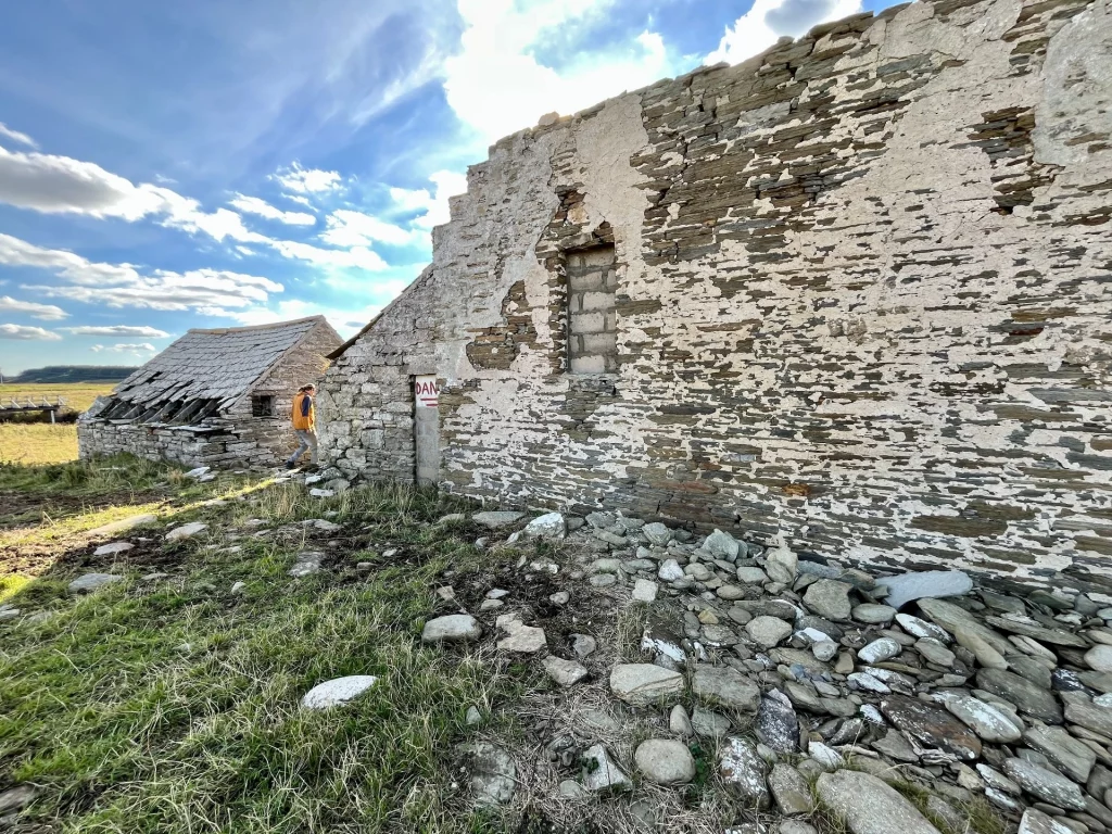 Weitwinkelaufnahme eines zerfallenen, flachen Steingebäudes unter blauem Himmel mit weißen Wolken. Der Boden davor ist mit losen Steinplatten und kargem Gras bedeckt.