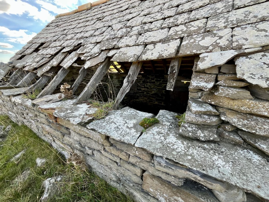 Nahaufnahme eines beschädigten Steindachs. Die Schieferplatten sind teilweise weggebrochen, wodurch die darunterliegenden, verwitterten Holzbalken und das dunkle Innere des Gebäudes sichtbar werden.