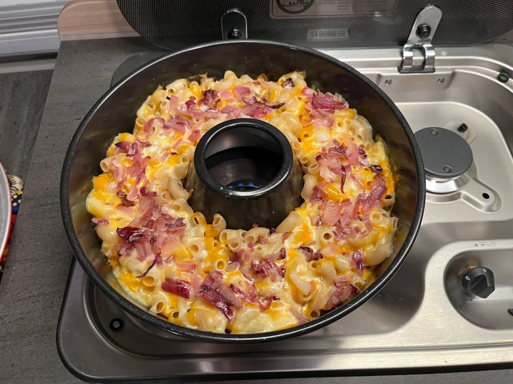 Draufsicht auf eine runde Omnia-Backform, die mit Kässpätzle, geschmolzenem Käse und reichlich Röstzwiebeln gefüllt ist und auf einem Gasherd im Wohnmobil steht.