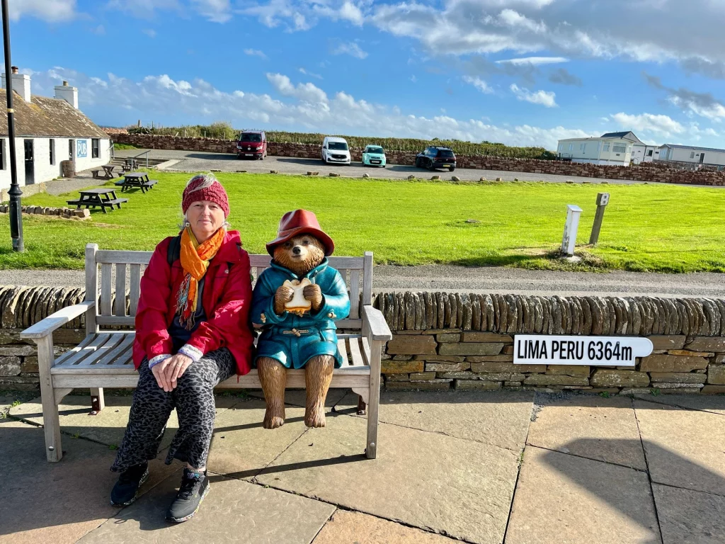 Karin sitzt neben einer Paddington-Bär-Statue am Hafen von John o' Groats vor dem Schild nach Lima, Peru.