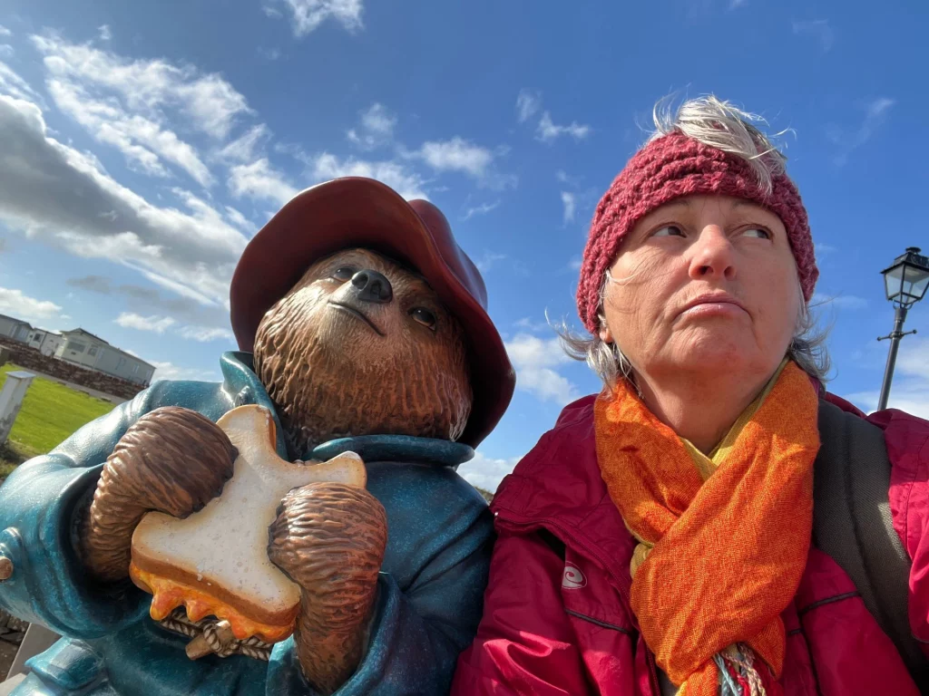 Karin sitzt neben einer Paddington-Bär-Statue am Hafen von John o' Groats vor dem Schild nach Lima, Peru.