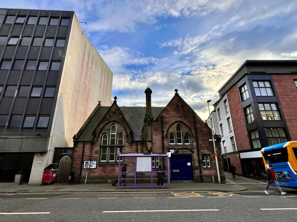 Ein historisches, kirchenähnliches Steingebäude mit spitzen Giebeln und gotischen Fenstern, das zwischen zwei modernen, höheren Funktionsbauten an einer Straße mit Bushaltestelle steht.