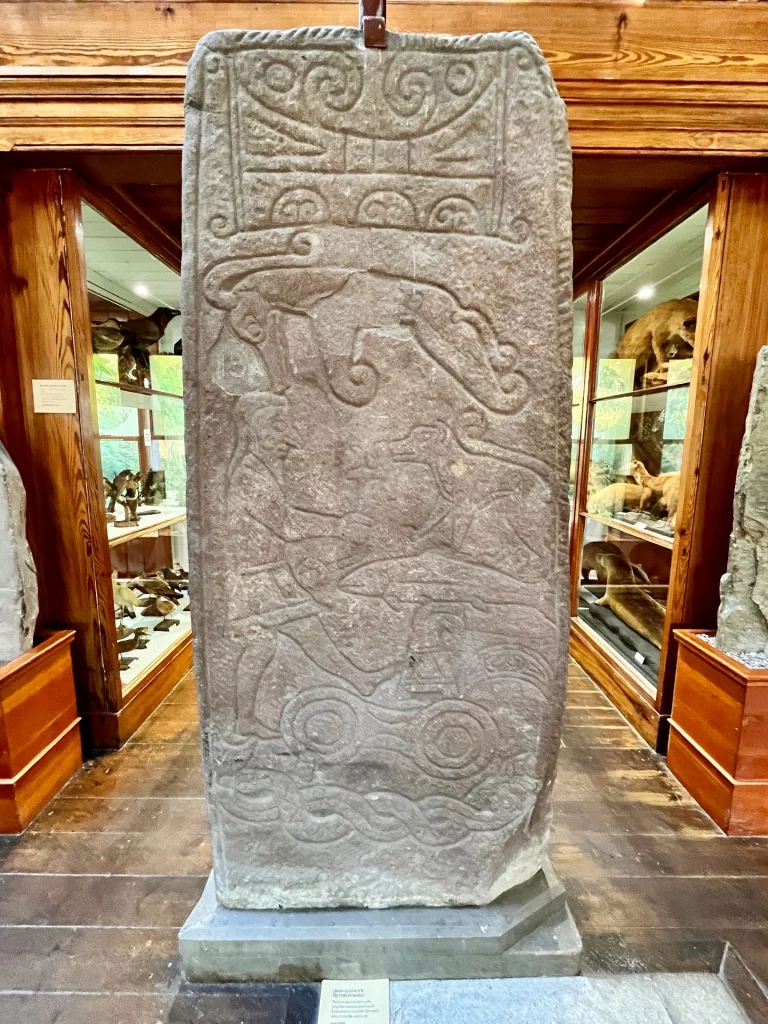 Eine hohe Steinplatte mit Reliefs von Tieren, einer menschlichen Figur und komplexen Knotenmustern.