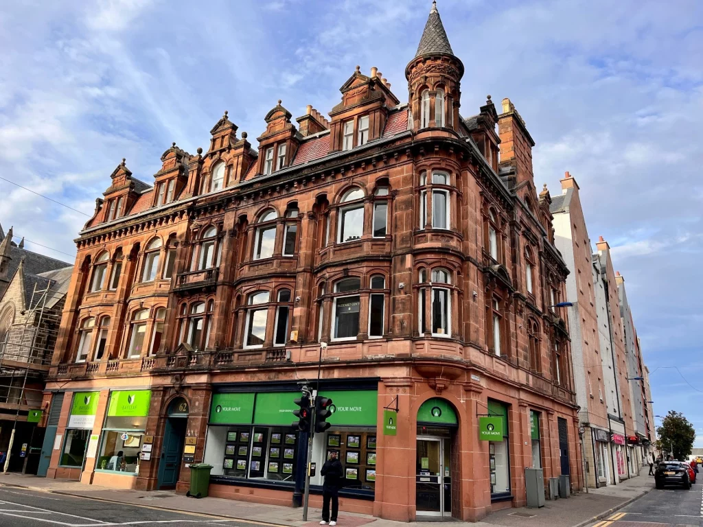 Ein mehrstöckiges, herrschaftliches Gebäude aus rotem Sandstein mit aufwendigen Verzierungen, Giebeln und einem kleinen Ecktürmchen unter einem leicht bewölkten blauen Himmel.