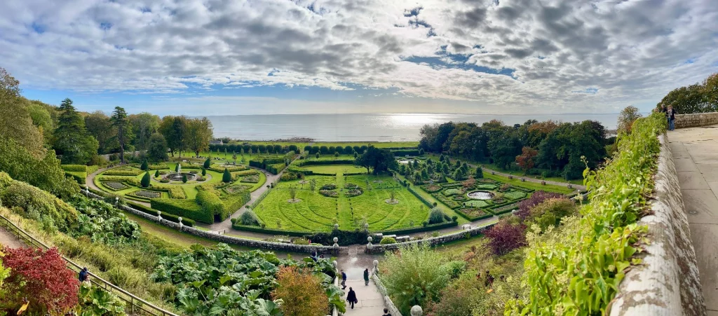 Eine Panorama-Aufnahme des weitläufigen, symmetrisch angelegten Barockgartens von Dunrobin Castle mit gepflegten Rasenflächen, Springbrunnen und Heckenlabyrinthen. Im Hintergrund grenzt der Garten direkt an eine ruhige Meeresbucht unter einem leicht bewölkten Himmel. Die Perspektive ist von einem erhöhten Standpunkt aus aufgenommen.