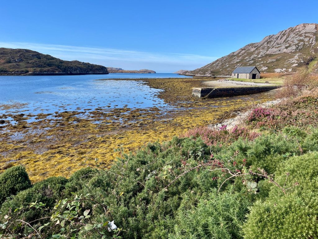 Ein kleines, einsames graues Steinhaus (Cottage) steht verlassen in der weiten, kargen Hügellandschaft der schottischen Highlands am Ufer eines Sees.