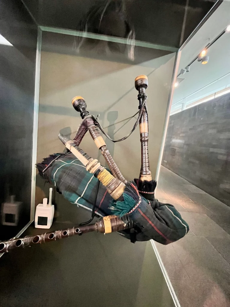 Ein alter schottischer Dudelsack aus dunklem Holz und grün-blauem Karostoff (Tartan), der in einer beleuchteten Glasvitrine im Culloden Besucherzentrum ausgestellt ist.