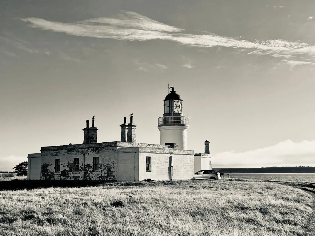 Schwarz-weiß Fotografie eines historischen Leuchtturms mit angrenzendem Wohnhaus an einer grasbewachsenen Küste unter einem weiten Himmel.