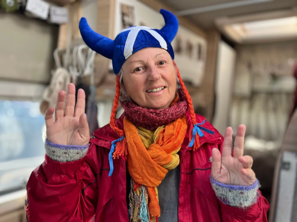 Porträt einer lächelnden Frau in einer roten Outdoorjacke im Inneren eines Wohnmobils. Sie trägt eine blaue Wikingerhaube mit weißen Hörnern und dem schottischen Andreaskreuz sowie einen bunten Schal.