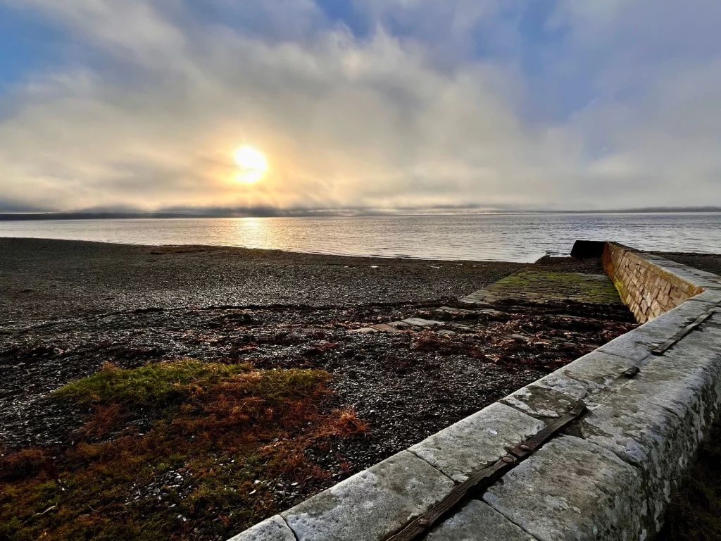 Goldener Sonnenaufgang am Chanonry Point in Schottland. Die Morgensonne bricht durch eine dramatische Wolkendecke über dem ruhigen Wasser des Moray Firth. Im Vordergrund eine historische Steinmauer am Kiesstrand.