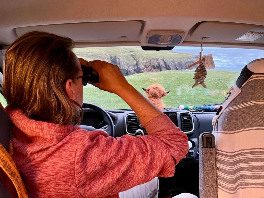 Innenansicht aus dem Fahrerhaus eines Campers; ein Mann (Ingo) blickt konzentriert durch ein Fernglas auf die grünen Klippen der Küste.
