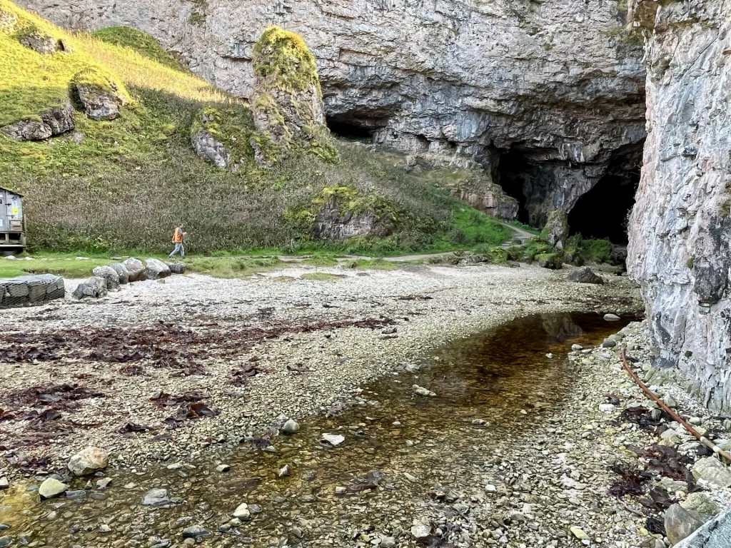 Eine weite Außenaufnahme der Smoo Cave in Durness, die den steinigen Vorplatz mit einem kleinen Bachlauf und die hohen Kalksteinklippen zeigt.