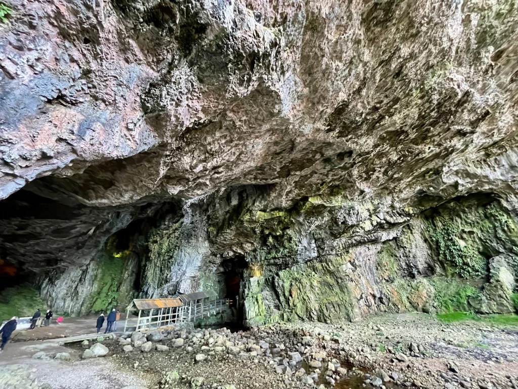 Detailansicht der massiven, teils moosbewachsenen Felswände im Eingangsbereich der Smoo Cave mit einer kleinen überdachten Brücke über dem Wasser.
