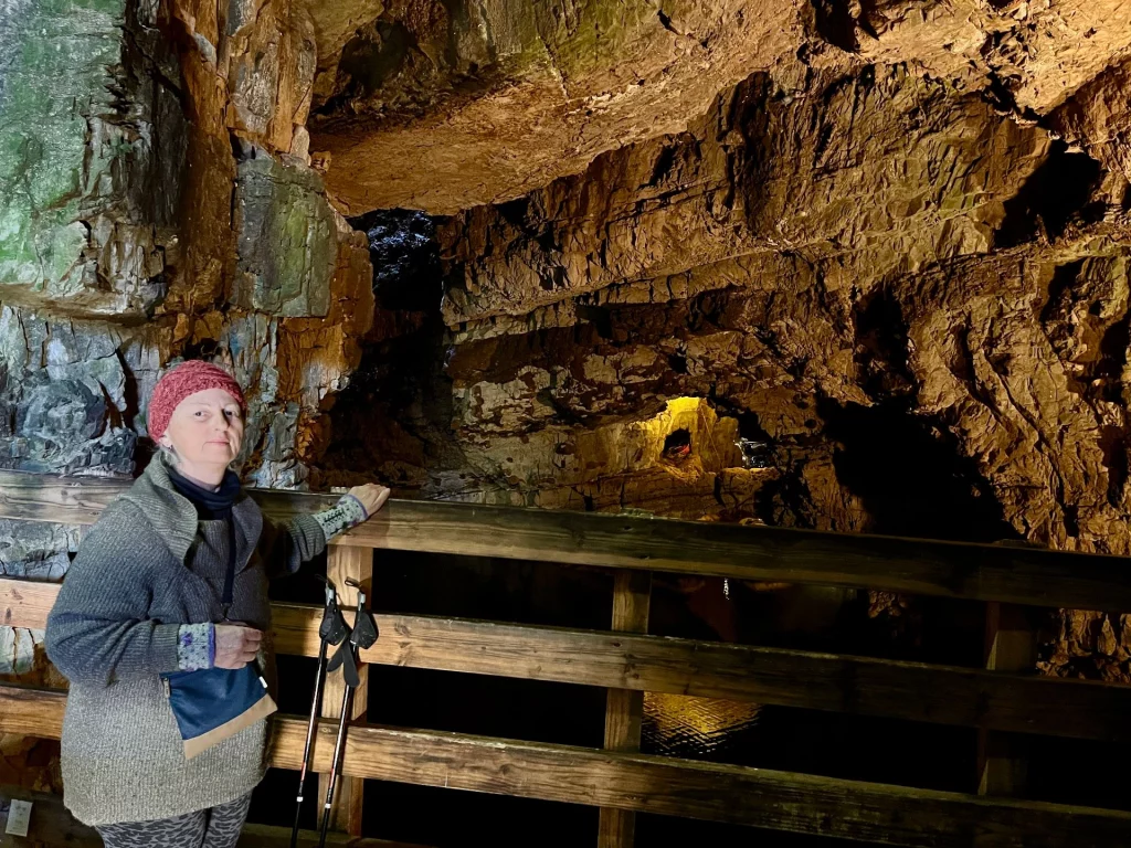 Eine Frau steht auf einem hölzernen Aussichtssteg in einer großen, von warmem Licht erleuchteten Felshöhle. Sie trägt eine rote Mütze, einen grauen Strickpullover und hat Wanderstöcke bei sich. Im Hintergrund sind die massiven, zerklüfteten Felswände der Höhle und eine kleine, hell erleuchtete Öffnung im hinteren Bereich zu sehen, die sich im dunklen Wasser darunter spiegelt.