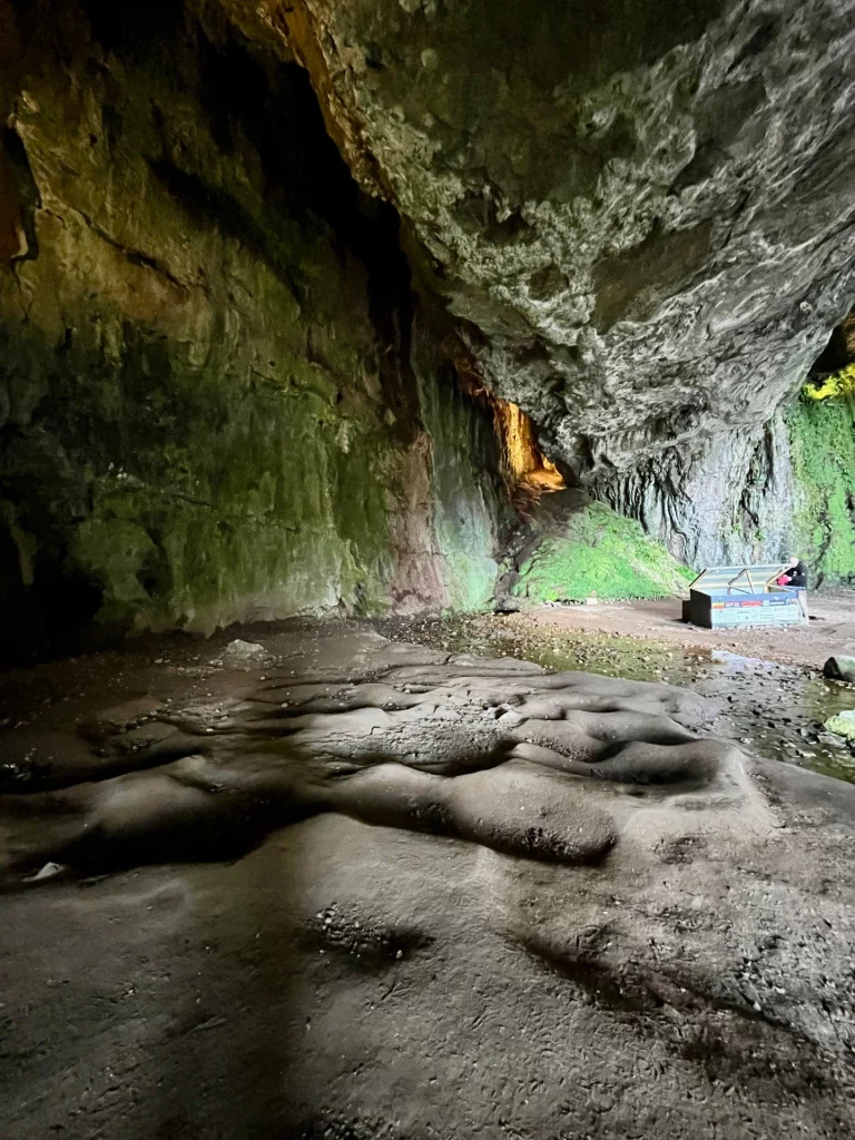 Eine weite Innenansicht der Smoo Cave mit sandigem, unebenem Boden. Das natürliche Licht fällt von rechts in die Höhle und beleuchtet eine kleine Informationshütte oder -tafel, an der eine Person steht. Die massiven Felswände zeigen grüne Moosakzente und führen im Hintergrund zu einem schmalen, hell erleuchteten Durchgang.