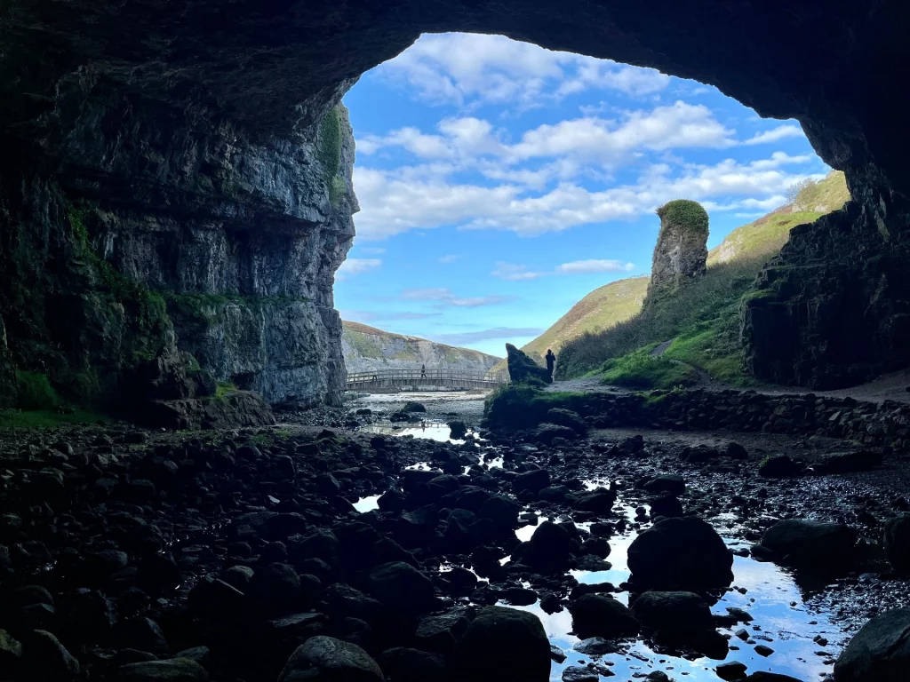 Der Blick aus dem riesigen, dunklen Höhlenportal der Smoo Cave nach draußen. Im Vordergrund liegen dunkle, nasse Steine in flachem Wasser, die den blauen Himmel reflektieren. Im hellen Bildzentrum sieht man eine Holzbrücke, grüne Grashügel und einen markanten Felsen unter einem leicht bewölkten Himmel.