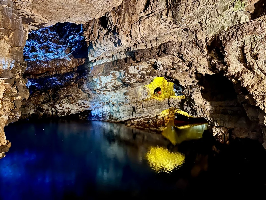Eine atmosphärische Aufnahme des unterirdischen Sees in der Smoo Cave. Das Wasser ist tiefblau und ruhig, darin spiegelt sich eine punktuelle, gelbe Beleuchtung der zerklüfteten Höhlendecke. Die Felsformationen wirken dramatisch und sind durch künstliches Licht in kontrastreichen Blau- und Goldtönen in Szene gesetzt.