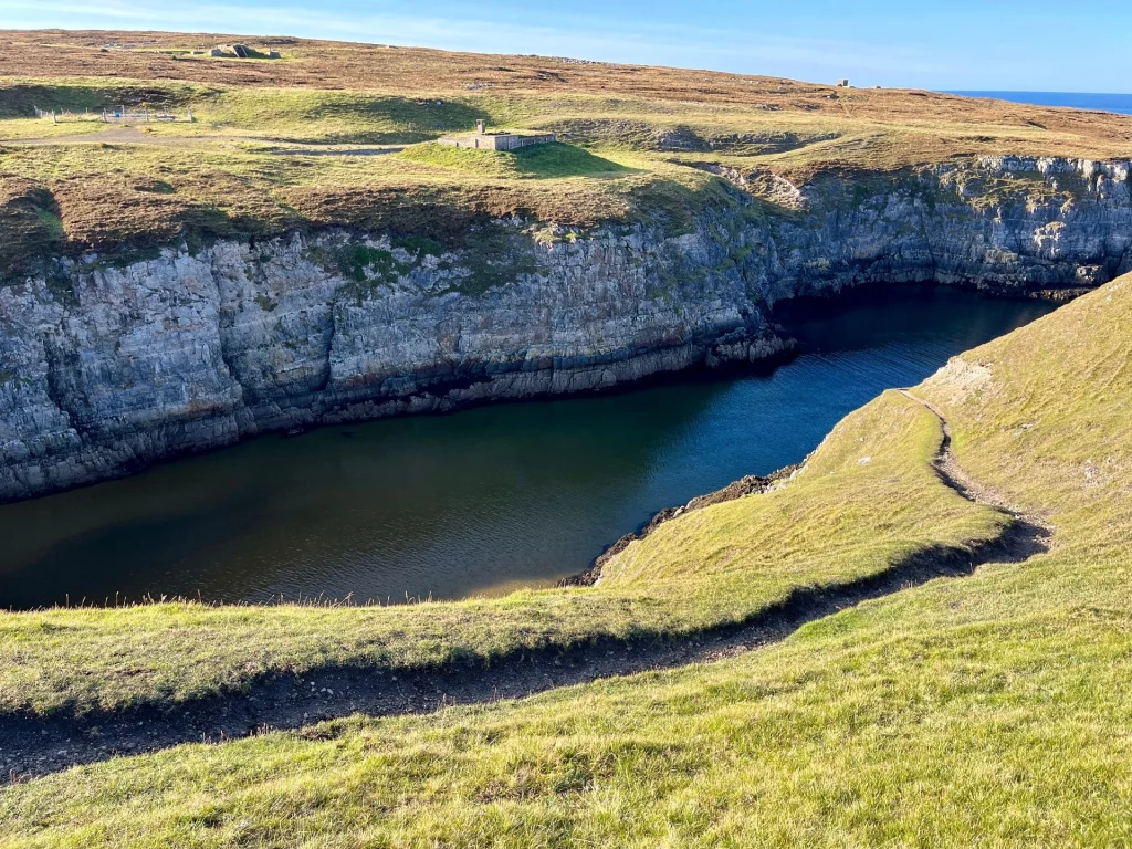 Smoo Cave Klippen Bucht: Blick von oben auf eine tief eingeschnittene Meeresbucht mit steilen, schroffen Felswänden. Ein schmaler Wanderpfad schlängelt sich am grasbewachsenen Hang entlang.