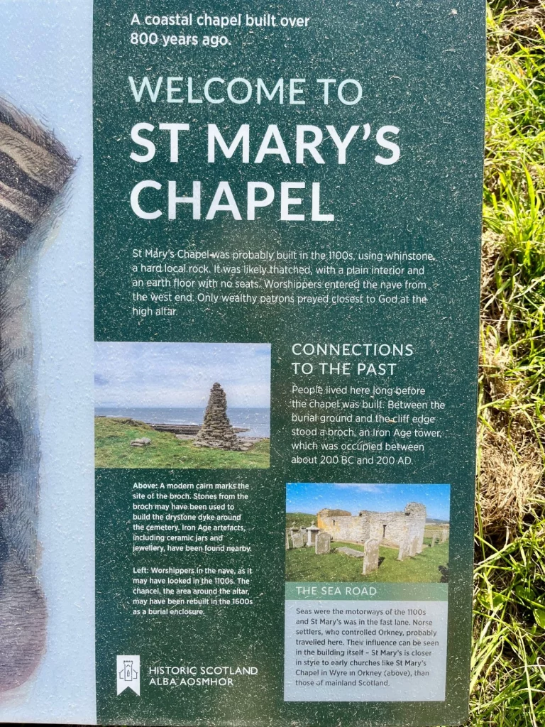 Eine grün-weiße Informationstafel von „Historic Scotland“. Der Text trägt die Überschrift „Welcome to St Mary’s Chapel“ und erklärt, dass die Kapelle über 800 Jahre alt ist. Kleine Begleitfotos auf der Tafel zeigen die Kapelle in der Küstenlandschaft.