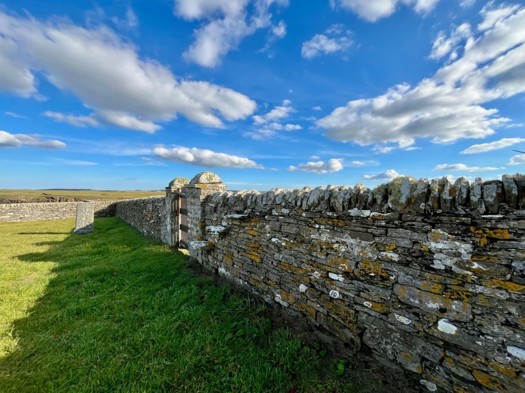 Eine lange, alte Trockenmauer aus grauem Stein zieht sich unter einem weiten, blau-weißen Wolkenhimmel durch die grüne Landschaft.