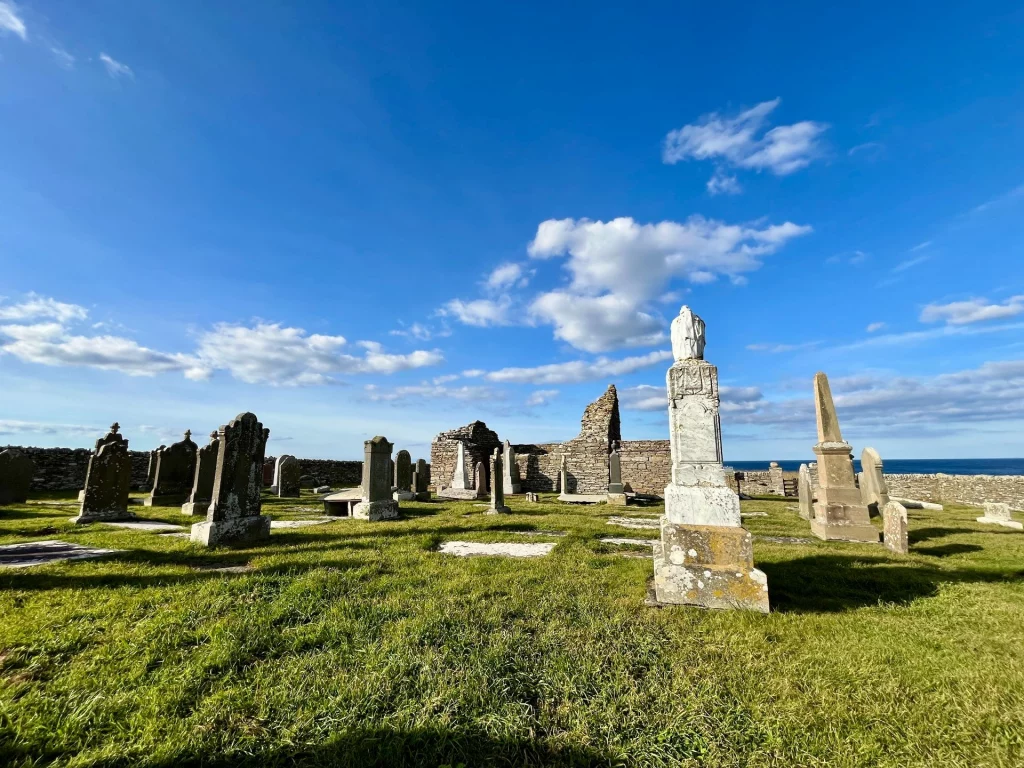 Weitwinkelaufnahme des Friedhofs mit verstreuten Grabsteinen unterschiedlicher Form und Größe. Im Hintergrund sind die Überreste der Kapelle und das Meer am Horizont erkennbar.