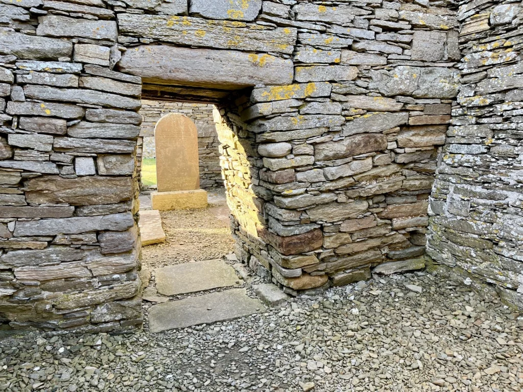 Nahaufnahme eines Torbogens: Eine massive Wand aus Trockenmauerwerk mit gelblichen Flechten umschließt einen rechteckigen Durchgang ohne Tür. Der Blick führt von einem kiesbedeckten Boden im Vordergrund auf eine sonnenbeschienene Fläche im Inneren, wo ein aufrecht stehender Gedenkstein und flache Grabplatten zu sehen sind.