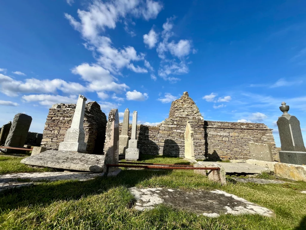 Eine beeindruckende Froschperspektive auf die Ruine der Kapelle. Im Vordergrund stehen verschiedene Monumente, darunter Obelisken und hohe Stelen, die sich gegen den strahlend blauen Himmel abzeichnen.