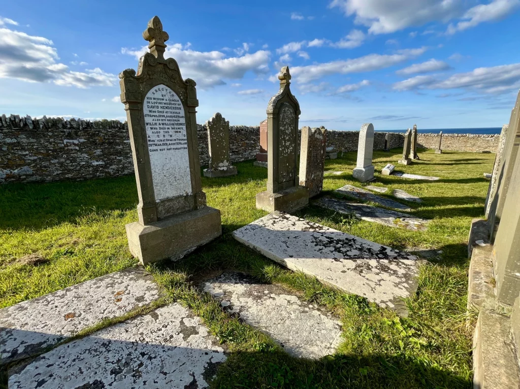 Mehrere kunstvoll verzierte, stehende Grabsteine auf einer grünen Rasenfläche. Die tiefstehende Sonne wirft lange Schatten über die im Boden eingelassenen Steinplatten.