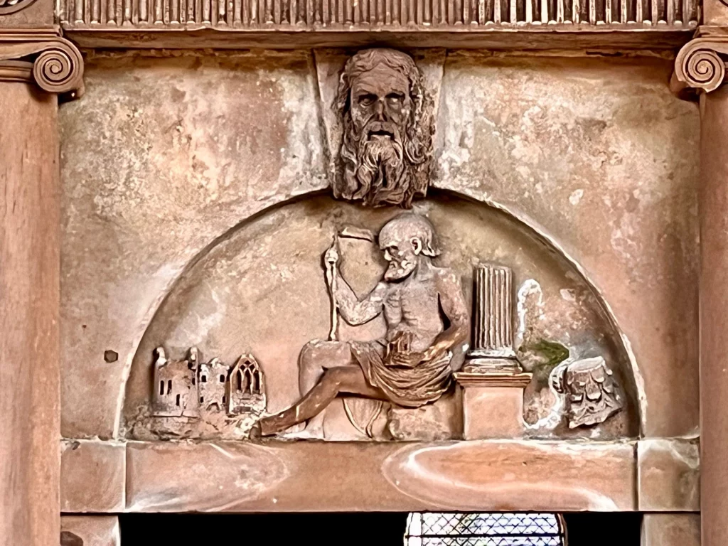 Detailaufnahme eines Steinfeldes (Tympanon) über einem Portal, das eine bärtige Figur mit einer Säule und Kirchenruinen zeigt.