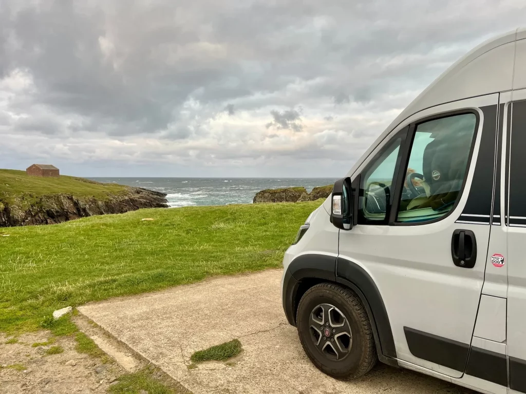 Seitenansicht eines weißen Kastenwagens, der auf einem ebenen Betonstellplatz direkt an der Abbruchkante einer grasbewachsenen Klippe mit Blick auf den weiten, grauen Atlantik steht.